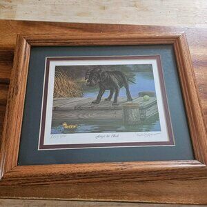 framed print of black Labrador retriever on a dock with a duck in water